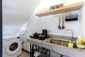 Une cuisine compacte équipée d’un lave-linge, d’un évier et d’un plan de travail sur lequel se trouvent une bouilloire, un grille-pain et une bouteille de liquide vaisselle.