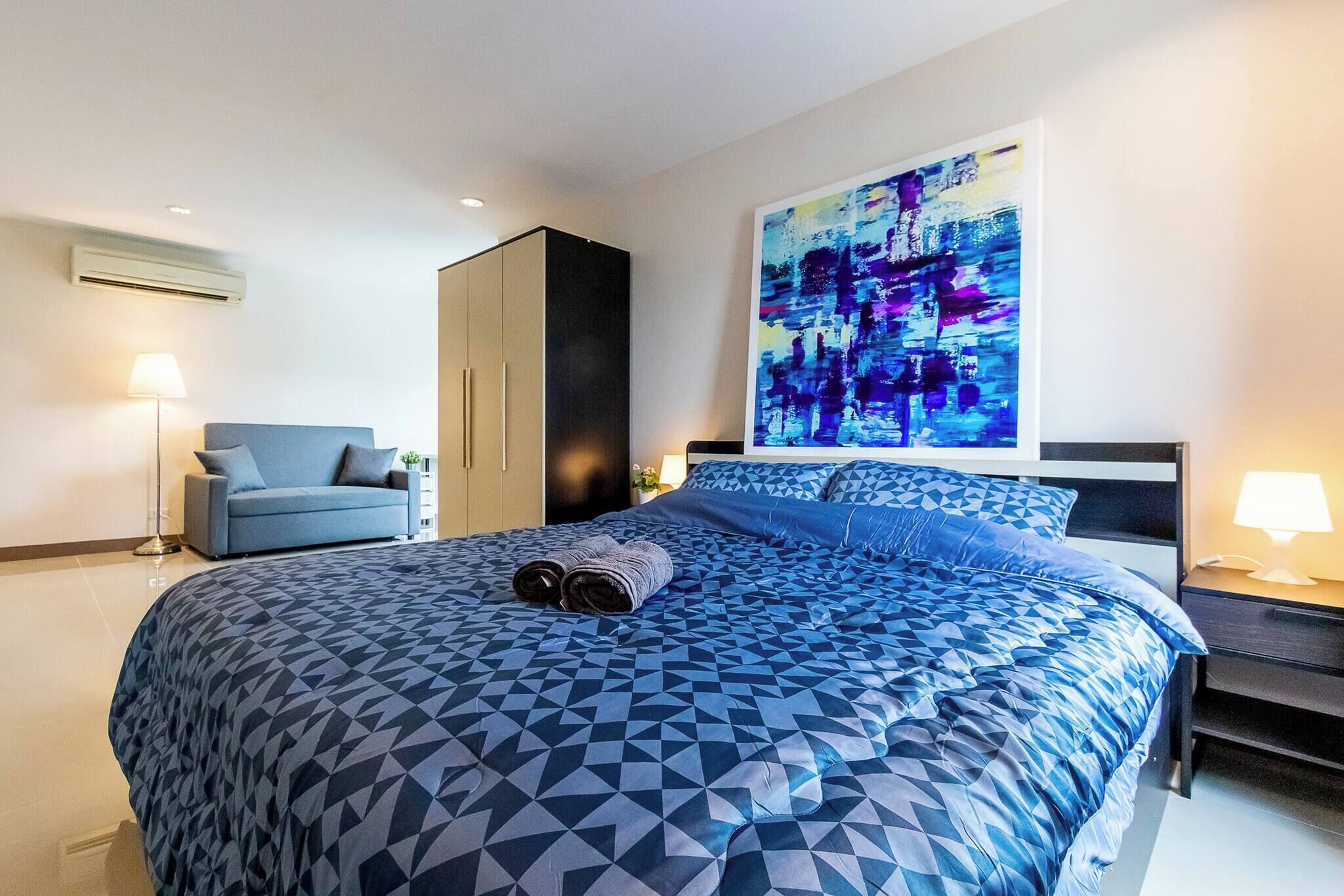Une chambre d’hôtel moderne avec un grand lit, une couvre-lit bleue à motifs géométriques, une grande peinture abstraite au mur, un fauteuil bleu et une table de chevet avec une lampe.