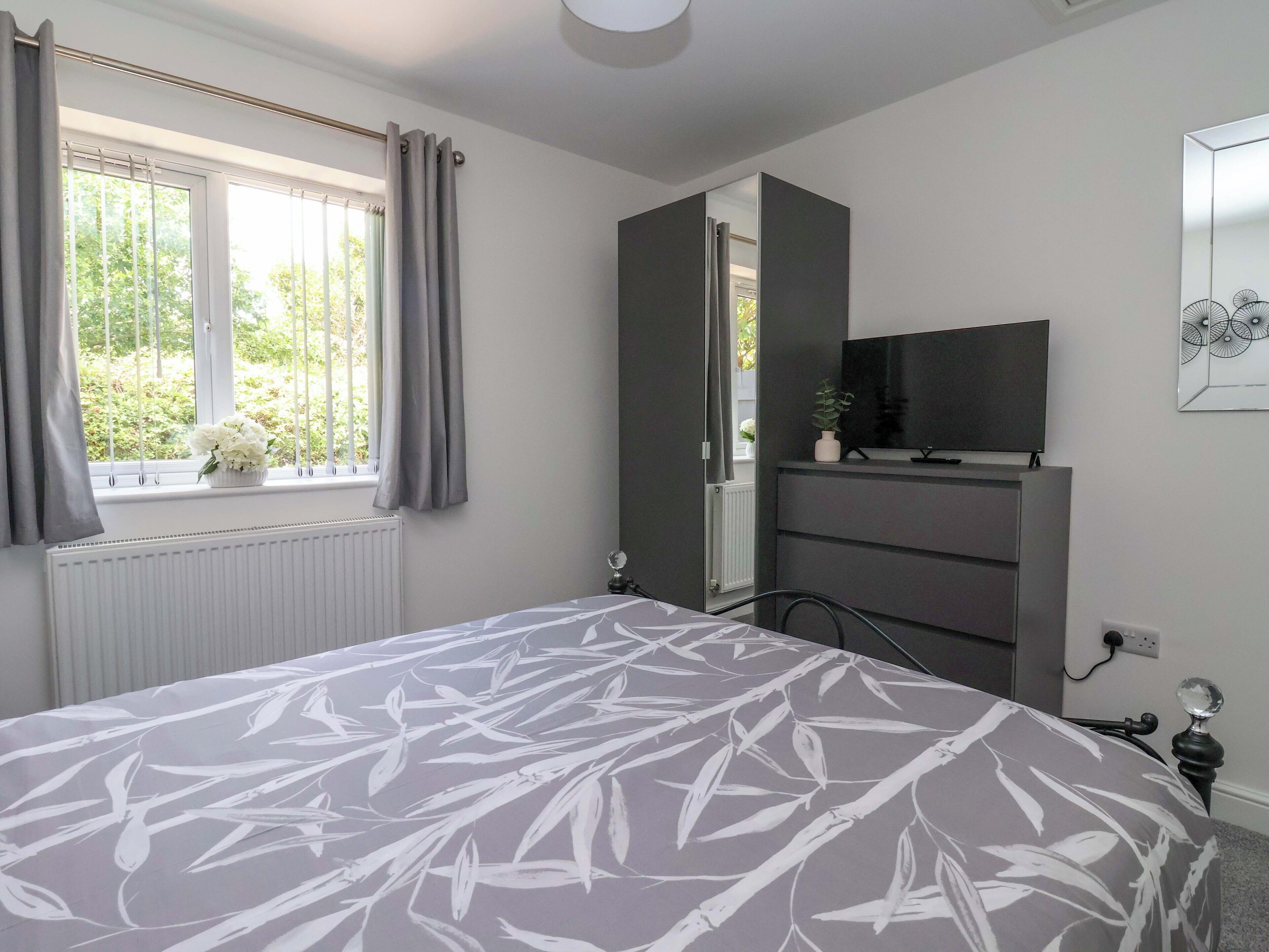 A bedroom with a bed, a window with curtains, a radiator, a TV on a cabinet, and a wall-mounted power outlet.