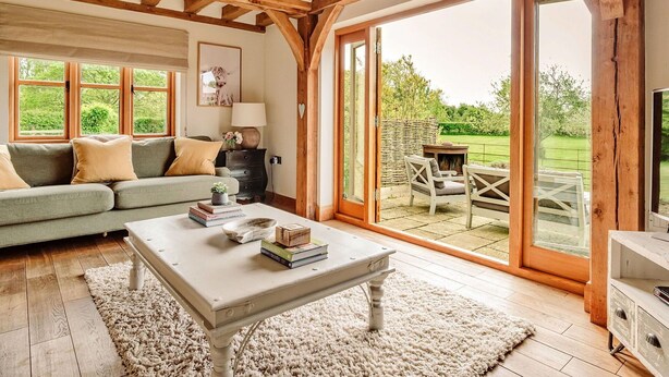 A cozy living room with a wooden ceiling, a sofa, a coffee table, and a view of a garden through large glass doors.