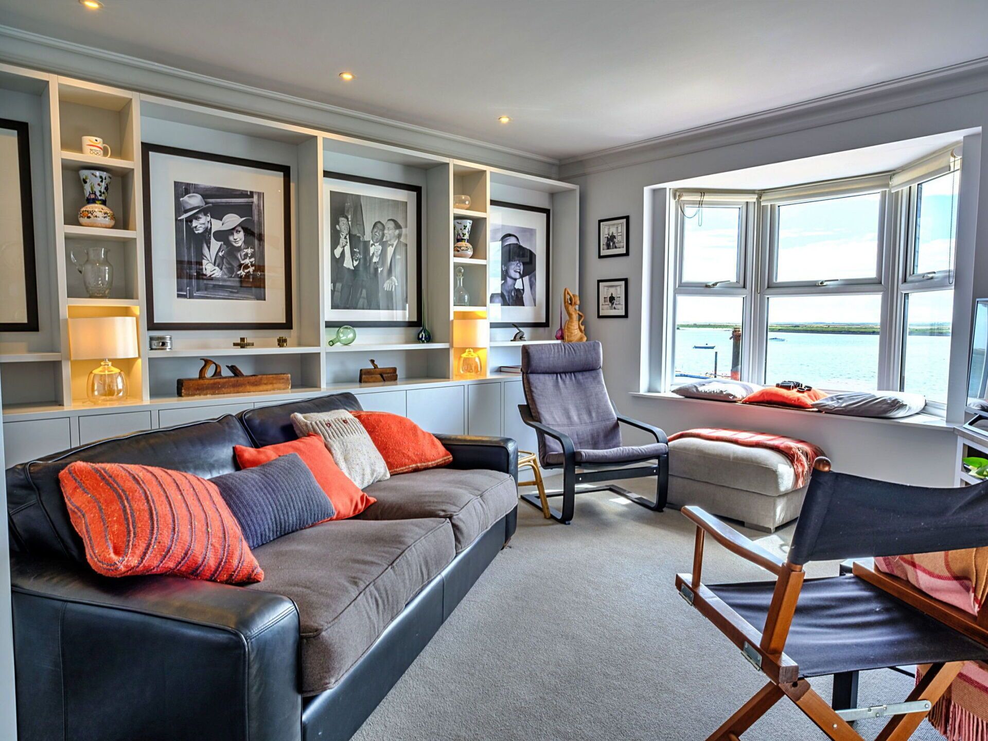 A modern living room with a grey sofa, a wooden chair, and a small table. There are shelves with decorative items and framed pictures on the wall.