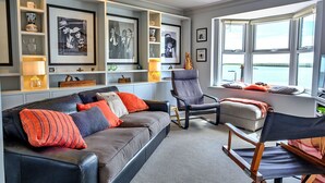Un salon moderne avec un canapé gris, une chaise en bois et une petite table. On y trouve des étagères ornées d’objets décoratifs et des tableaux encadrés accrochés au mur.