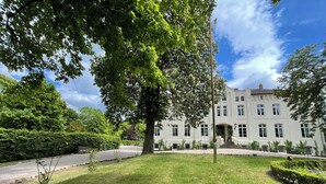 Een groot, wit gebouw met een symmetrische gevel, een centrale ingang met treden en een verzorgd gazon met bomen.