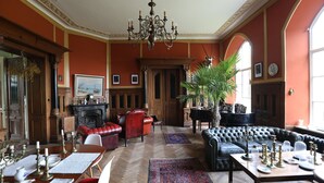 Een ruime kamer met een weelderige kroonluchter, een piano, een open haard en een groot raam.