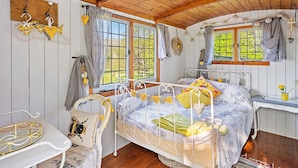A cozy bedroom with a wooden ceiling, two windows with curtains, a bed with floral bedding, a bedside table, and a chair with a bee design. - Stunning  villa for 4 people with pets allowed (Romsey)