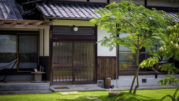 A traditional Japanese building with a tiled roof, a wooden door, and a small garden area with a tree and a hammock. - OMISHIMA SPACE HANARE (Imabari)