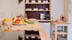A kitchen with wooden shelves holding white dishes and a coffee maker. There is a fruit bowl on the counter and a cutting board with bread.