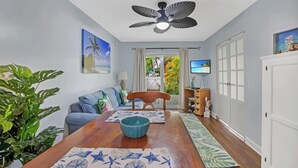 A living room with a wooden dining table, blue and white patterned mats, a ceiling fan, a TV, and a view of the outdoors through a glass door. - A SEAGLASS PARADISE ~ Charming 2 BR/1 Ba 1st Floor Condo in Truman Annex! (Key West)