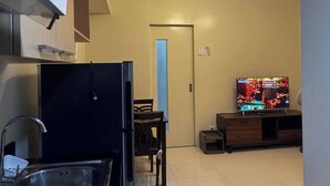A compact kitchen with a refrigerator, sink, and a dining area with a chair. A television is mounted on a wooden cabinet, displaying a music video. - Cozy Brenthill Condo in Baguio City - Entire House Rental (Baguio)