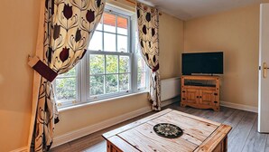 A room with a large window, a wooden cabinet with a flat-screen TV, and a round wooden table with a decorative plate.