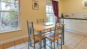 A dining area with a wooden table and metal chairs, a kitchen with white cabinets, and large windows with curtains.