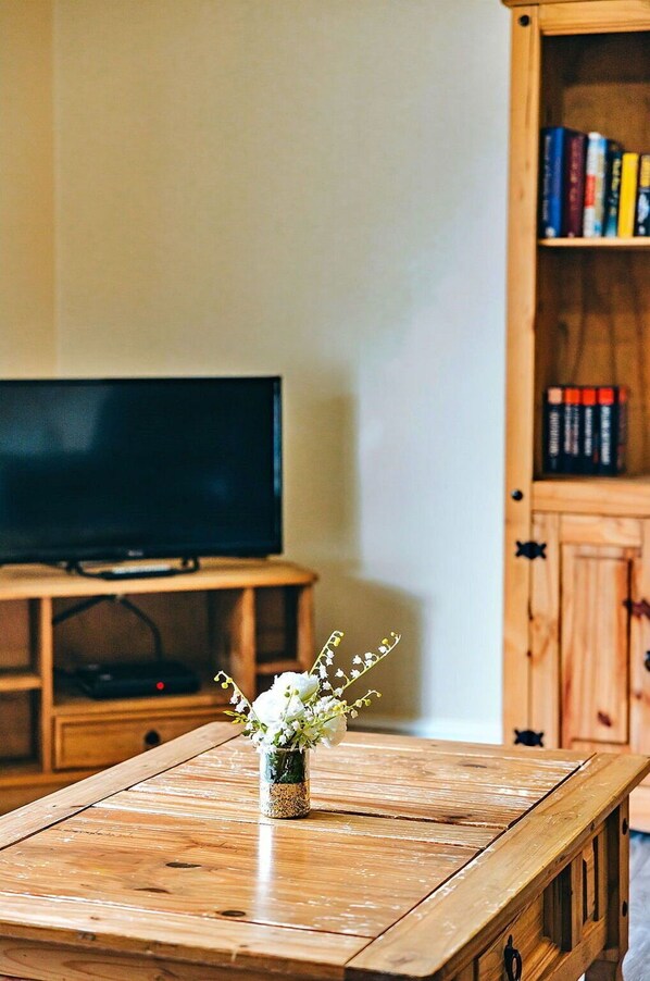 A wooden table with a vase of flowers in the foreground, a television on a stand in the background, and a wooden bookshelf with books. - Stunning  villa for 2 people with WIFI, TV and pets allowed (Shrewsbury)