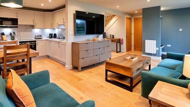 A modern kitchen with white cabinets, a built-in oven, and a large island with a sink. There is a dining area with a wooden table and chairs, and a living room with a green sofa and a coffee table.