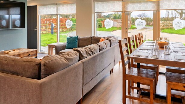 A modern living room with a sectional sofa, a dining table set for six, and large windows offering a view of a garden. - Beautiful  villa for 8 people with WIFI, TV and pets allowed (Alcester)