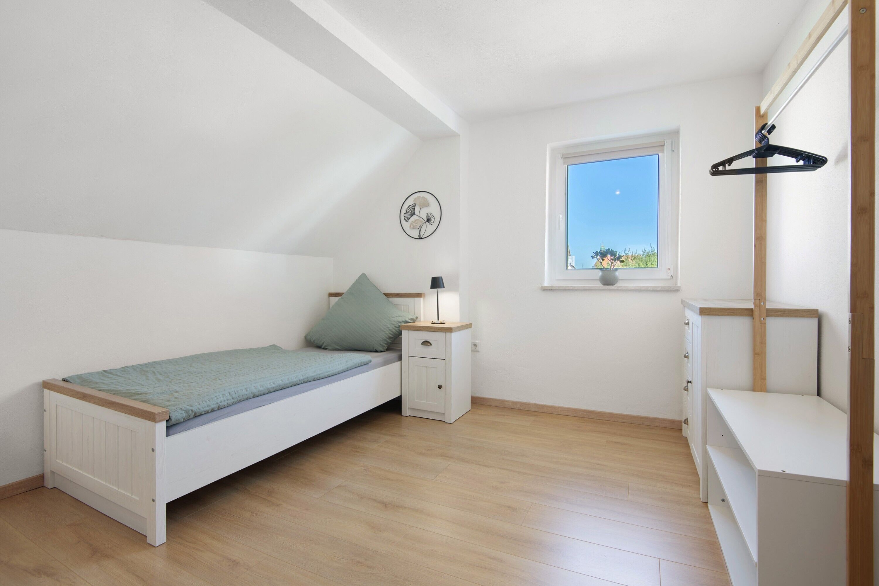Een compacte, goed verlichte kamer met een enkel bed, een nachtkastje, een dressoir, een raam en een ventilator bevestigd aan de muur.