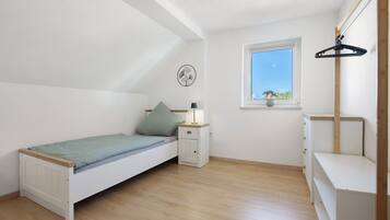 Een compacte, goed verlichte kamer met een enkel bed, een nachtkastje, een dressoir, een raam en een ventilator bevestigd aan de muur.