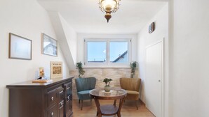 Una habitación con suelo de madera, una mesa redonda con sillas, un mueble de madera oscura y dos ventanas con vistas a un edificio.