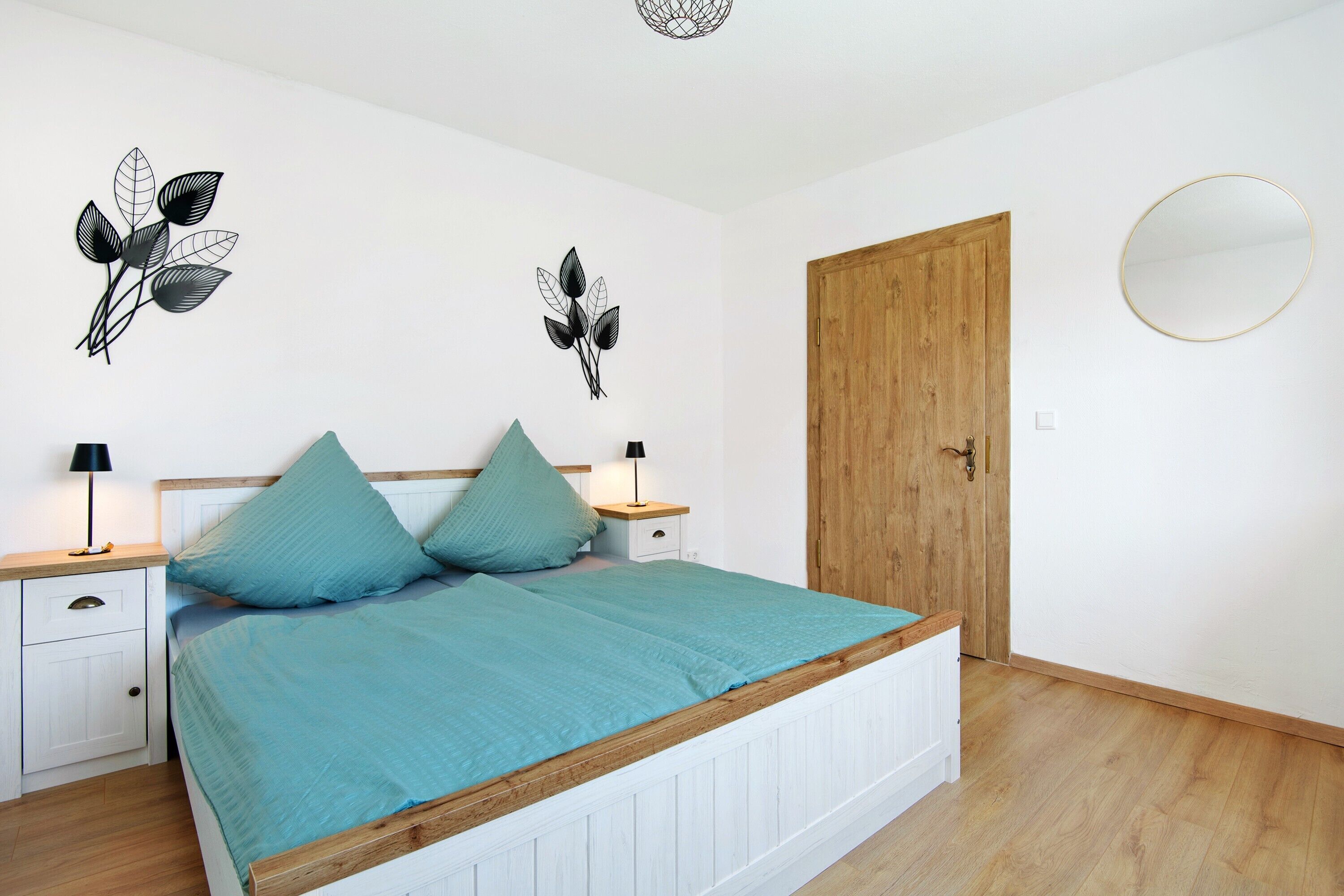 Une chambre à coucher comprenant un lit, deux tables de chevet, une porte en bois, un miroir rond et des œuvres d’art murales.