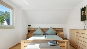 Un dormitorio con cama, banca, mesitas de noche, una lámpara y un cuadro en la pared.