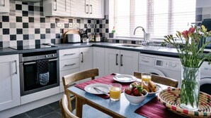 Een moderne keuken met witte kasten, een zwart-wit betegelde muur, een eettafel met ontbijtspullen en een raam met zonwering.