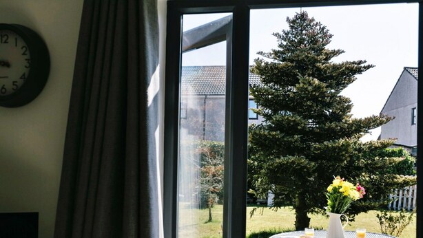 Een kamer met een groot raam, een klok aan de muur en uitzicht op een tuin met een tafel en stoelen.