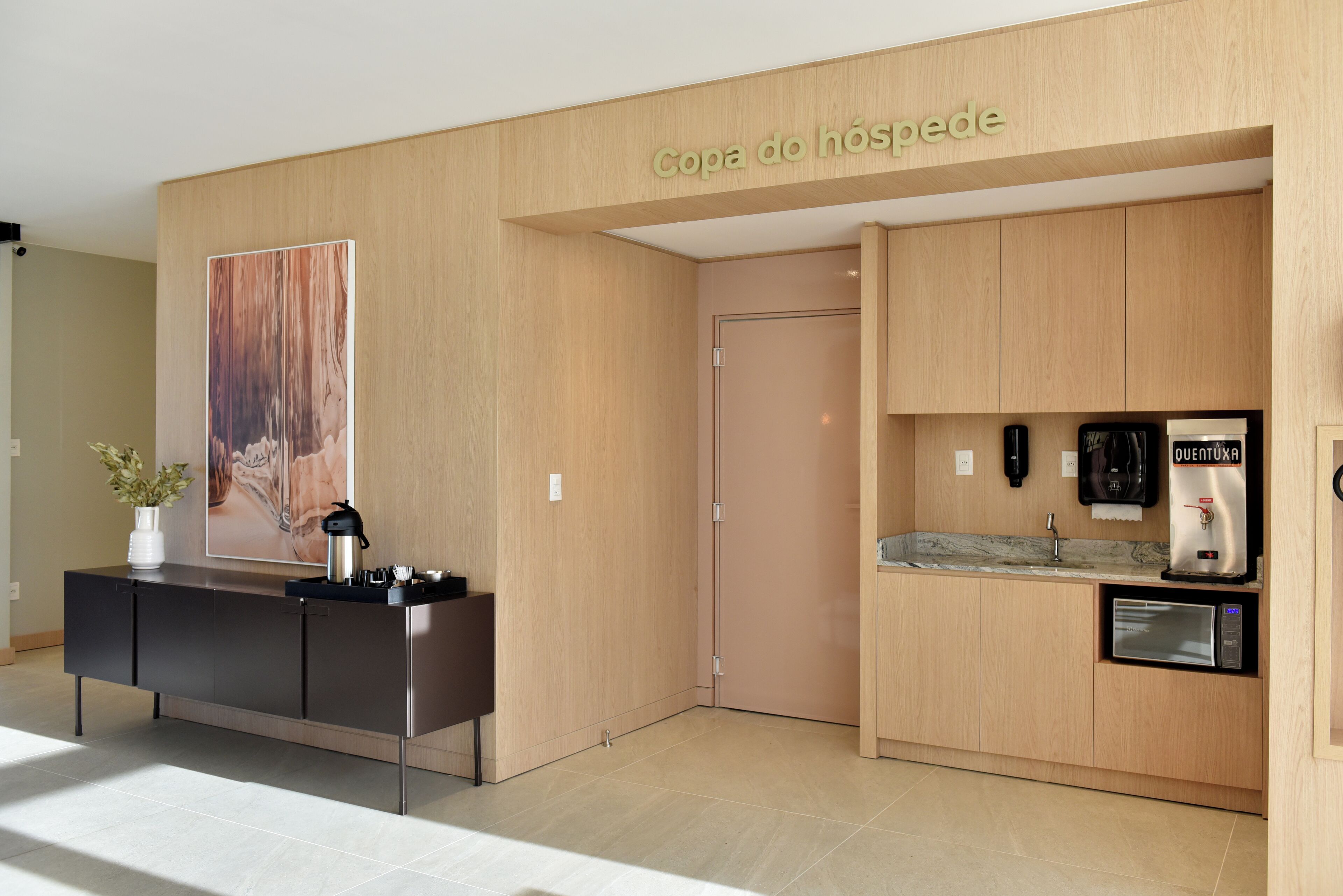 A reception desk with a coffee machine, a wall-mounted sign reading 'Copa do hóspede,' and a kitchen area with a coffee machine and a refrigerator.