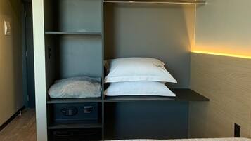 A compact hotel room with a bed, a built-in shelf unit, a wardrobe, and a wall-mounted air conditioning unit.