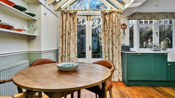 A dining area with a round wooden table, chairs, a bowl, a window with curtains, and a green kitchen with a sink.
