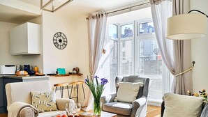 Una sala acogedora con un sofá, sillones, una mesa de centro con frutas y vasos, una mesita auxiliar y vistas de la ciudad a través de grandes ventanales.
