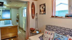 A cozy bedroom with a wooden bed, a small bedside table, a wall-mounted surfboard, and a window with curtains. - Stunning  villa for 2 guests with WIFI and terrace (Tintagel)