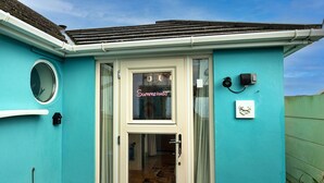 A turquoise house with a white-framed door, a small circular window, and a visible garden area with plants and a blue cover. - Stunning  villa for 2 guests with WIFI and terrace (Tintagel)