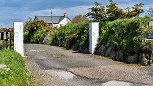 Un chemin de campagne mène à une maison blanche au toit vert, entourée de verdure et d’un mur de pierres.