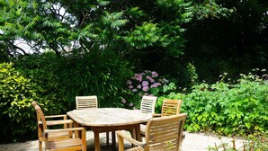 Une table et des chaises en bois sont disposées à l’extérieur, avec un jardin en arrière-plan.