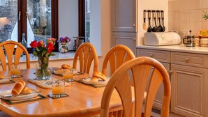 Een eethoek met een houten tafel, gedekt voor zes personen, omringd door houten stoelen. Op de tafel staat een vaas met bloemen en er is een raam met uitzicht op gebouwen en bomen.