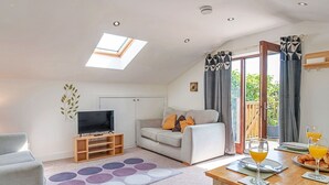 A modern living room with a flat-screen TV, grey sofa, and a dining table with breakfast items. - Lovely apartment for 3 people with terrace (Bodmin)
