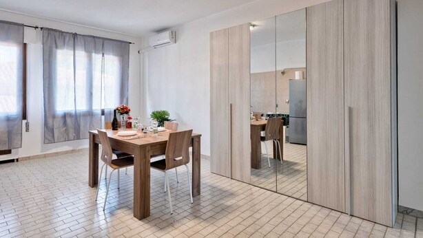 A modern dining area with a wooden table and chairs, a kitchen visible through a glass partition, and a large mirror reflecting the room. - Cozy Apartment 2 steps from the Railway Station (Chirignago-Zelarino)