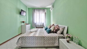 A bedroom with a bed, a TV on the wall, a window with curtains, and a bedside table with a lamp and water bottles. - Cozy Apartment 2 steps from the Railway Station (Chirignago-Zelarino)