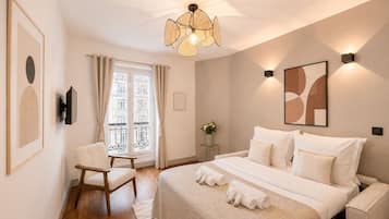 Un dormitorio con cama, una silla, una mesita de noche con un florero y una ventana con cortinas.
