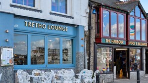 Un edificio con un letrero que dice "TRETHO LOUNGE" y otro letrero que anuncia "THE HARBOUR POOL & BILLIARDS". Hay sillas y mesas blancas al aire libre frente al edificio.
