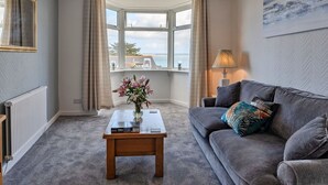A cozy living room with a grey sofa, a coffee table with a vase of flowers, a lamp, and a view of the sea through a large window. - Beautiful apartment for 4 guests with WIFI (St Ives)
