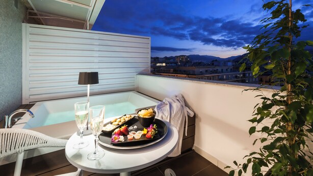 Uma varanda com mesa posta para refeição, uma garrafa de champanhe e vista para a cidade ao entardecer.