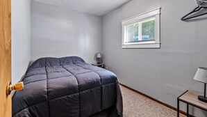 A bedroom with a bed, a nightstand, a lamp, a window with blinds, and a door.