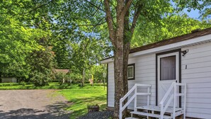 A small white cabin with a porch and steps, surrounded by lush green trees and a well-maintained lawn.