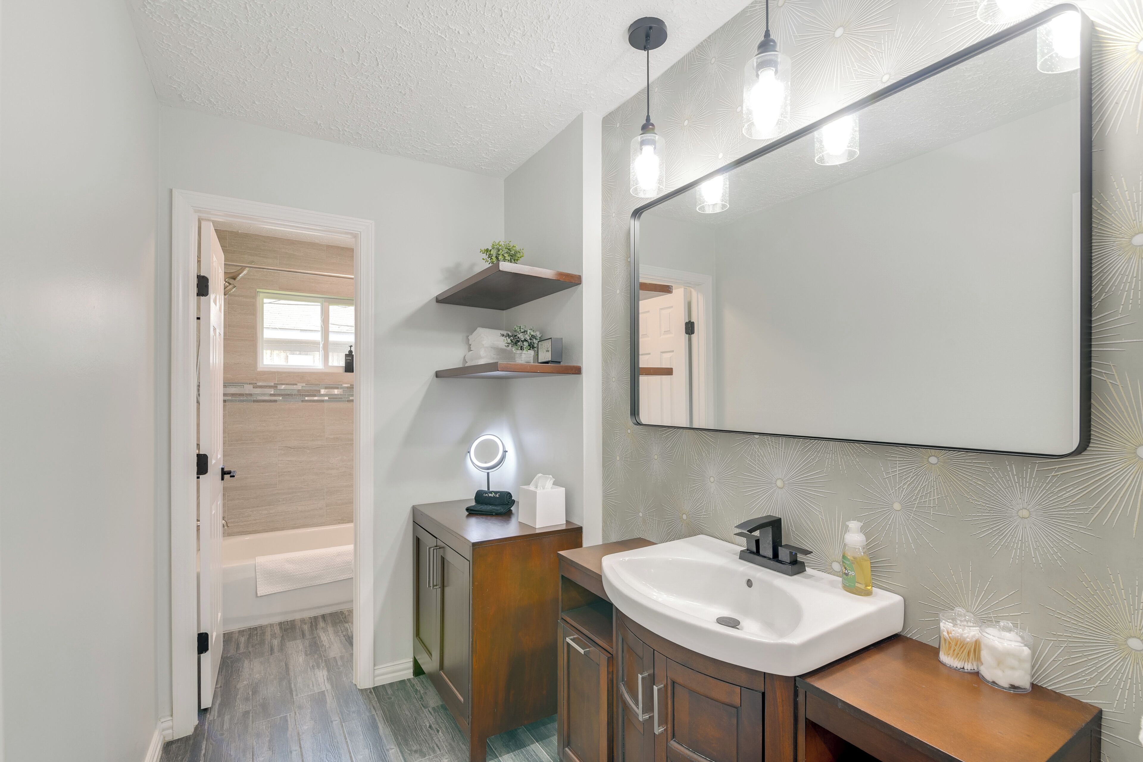 A bathroom with a white sink, wooden vanity, and a large mirror. There is a shelf above the sink with a plant and a towel.