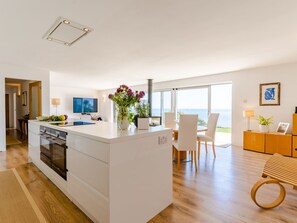 Cozinha moderna com ilha central, área de jantar com cadeiras e vista para o mar.