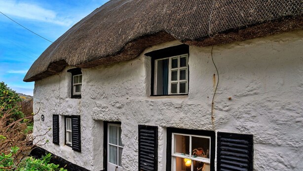 Une maison de campagne traditionnelle à toit de chaume, aux murs blancs et aux volets noirs, dotée d’un petit porche et d’une fenêtre visible, éclairée de l’intérieur.
