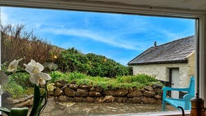 Une vue par une fenêtre, avec un mur de pierre, une chaise bleue et un bâtiment blanc en arrière-plan.