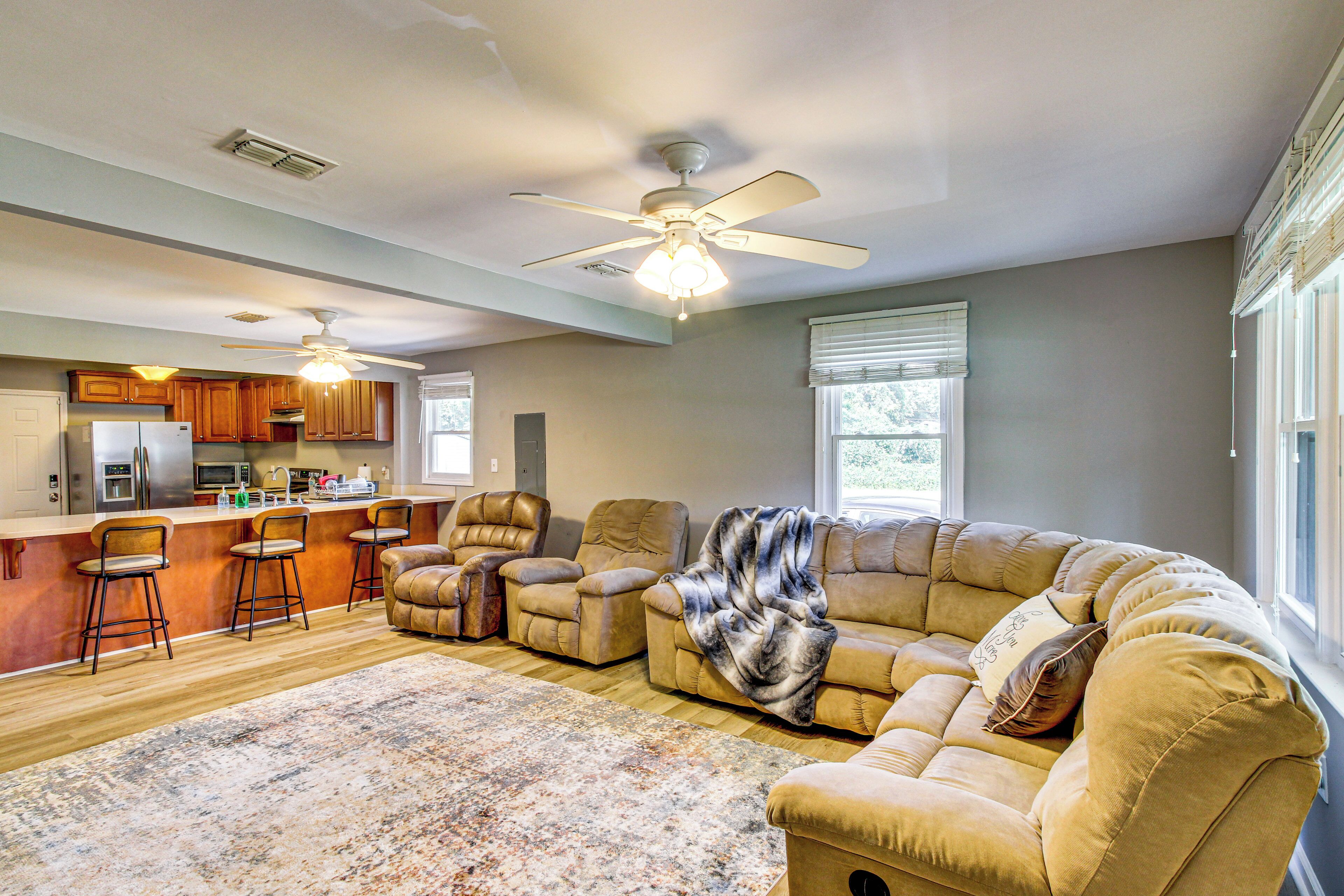 A spacious living room with a large beige sectional sofa, a kitchen area with wooden cabinets, and a ceiling fan.