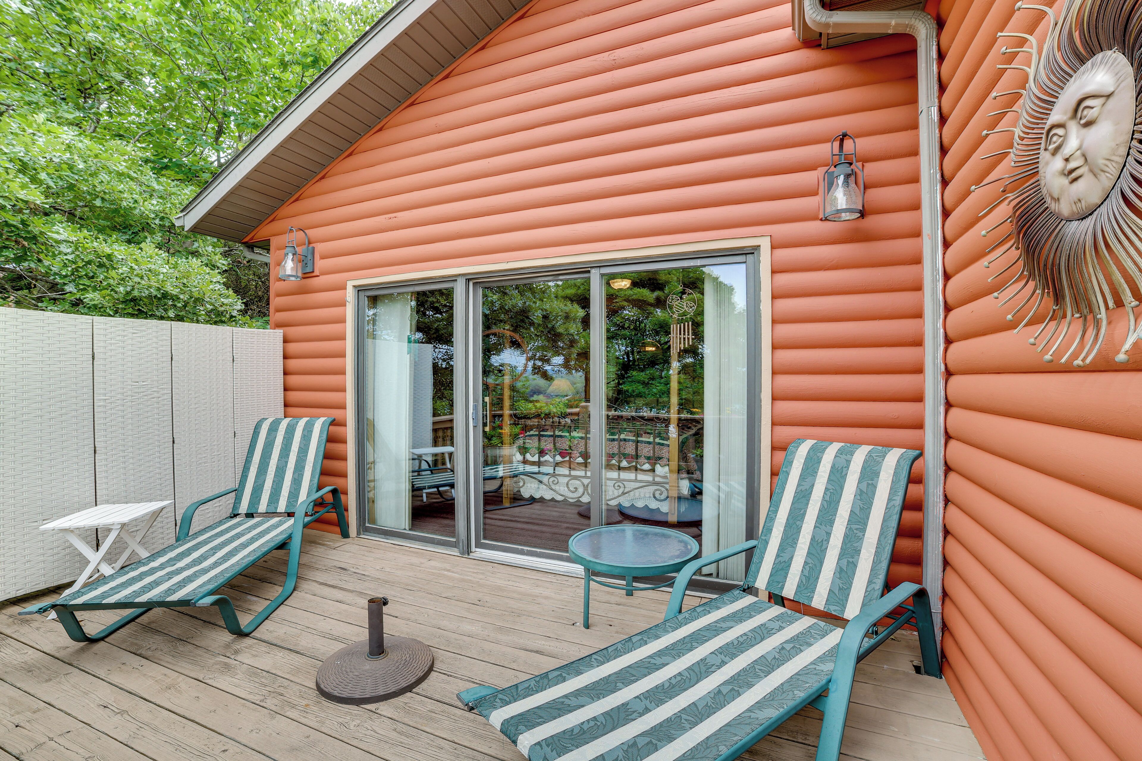 Une terrasse en bois avec deux chaises longues à rayures et une petite table, une porte vitrée donnant sur un espace extérieur, et une fresque murale décorative représentant un soleil.
