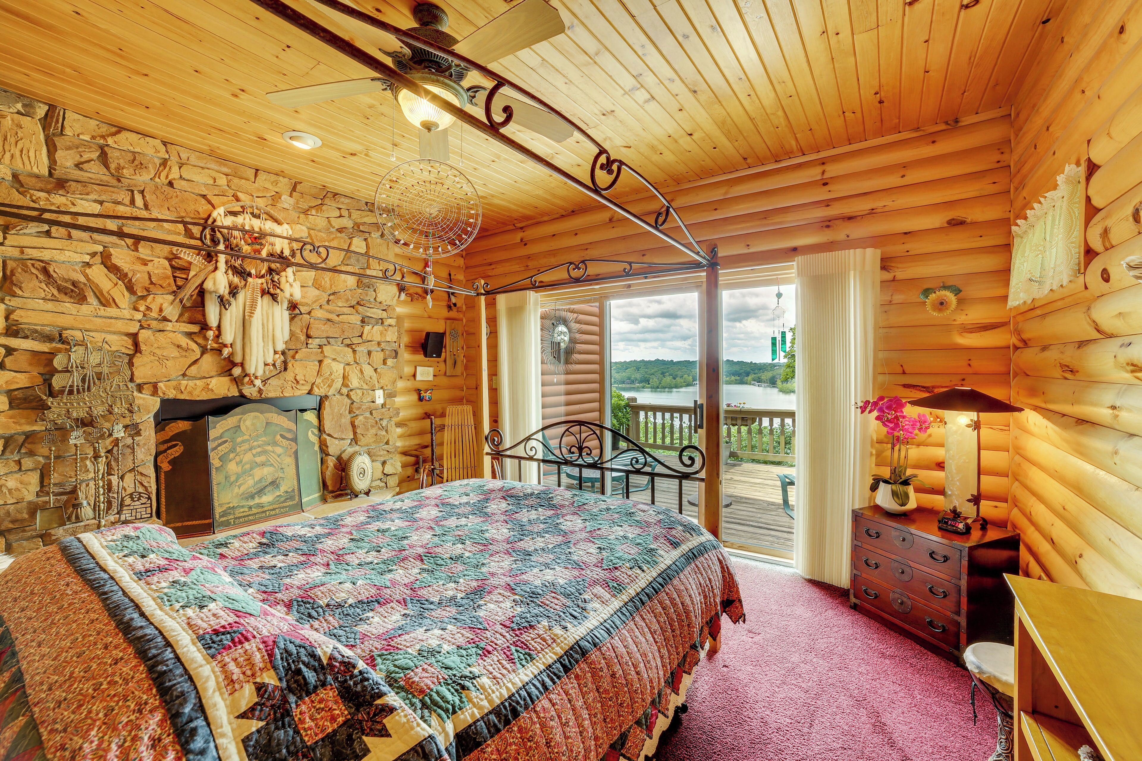 A log cabin bedroom with a large bed, stone wall, wooden ceiling, and a balcony with a view.